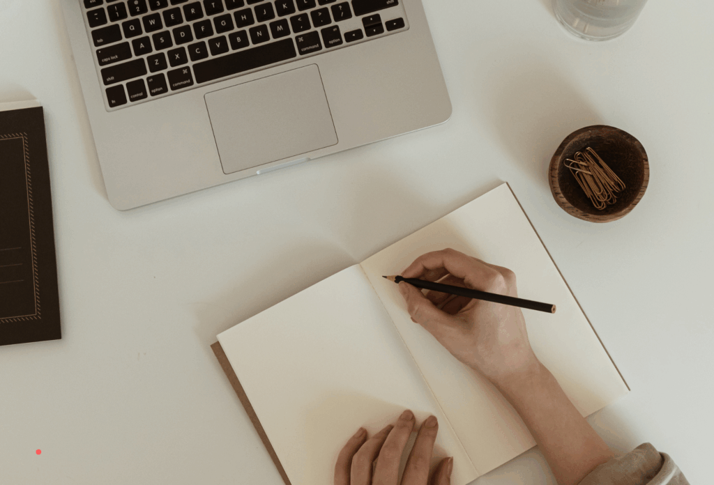 writing by hand in a notebook beside a laptop on a clean desk