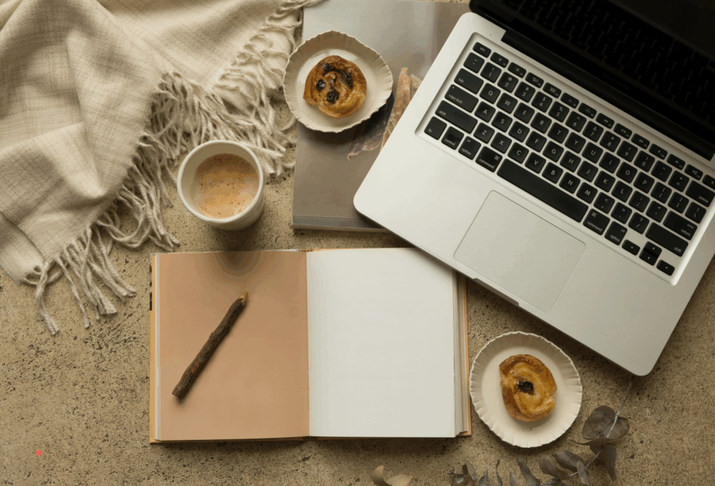 Cozy writing setup with an open notebook, laptop, coffee and pastries on a textured surface