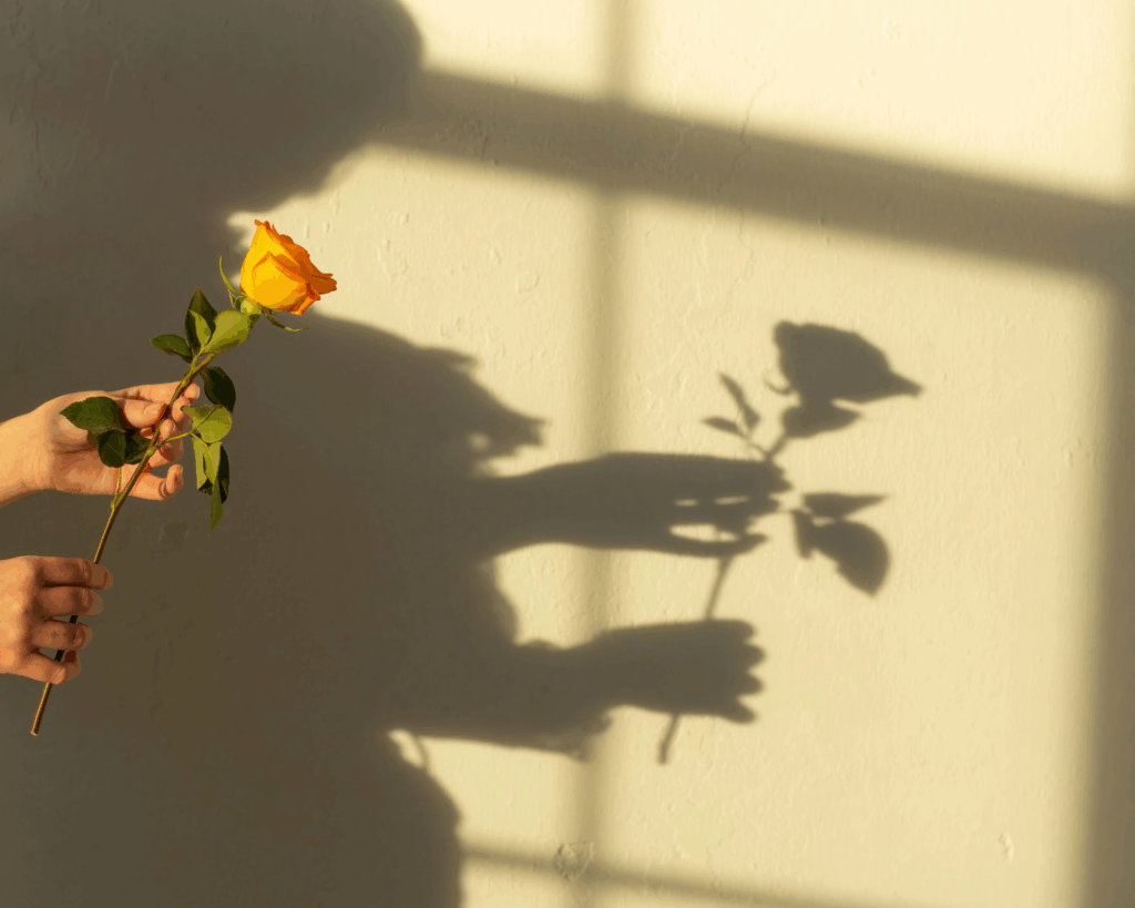 Hand holding a yellow rose against a wall with soft shadows, symbolizing offering, vulnerability, and quiet emotional presence.