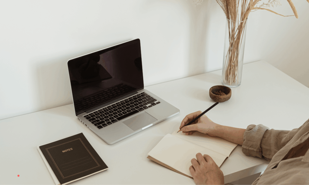 Person journaling by hand at a desk with a laptop, blending digital and handwritten reflection.