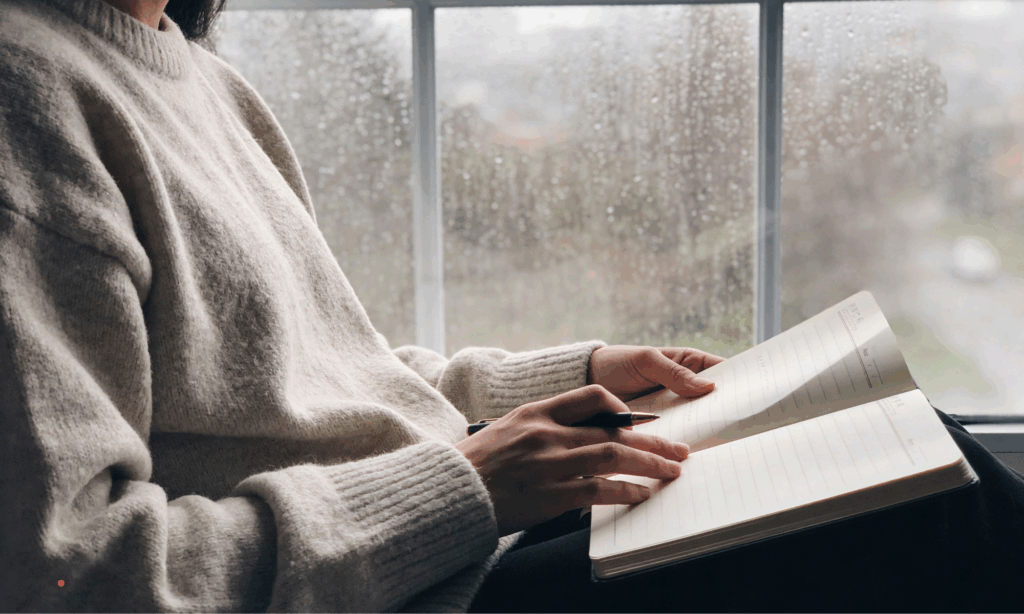 Person writing in a notebook by a rainy window, capturing a quiet moment of reflection and inner silence